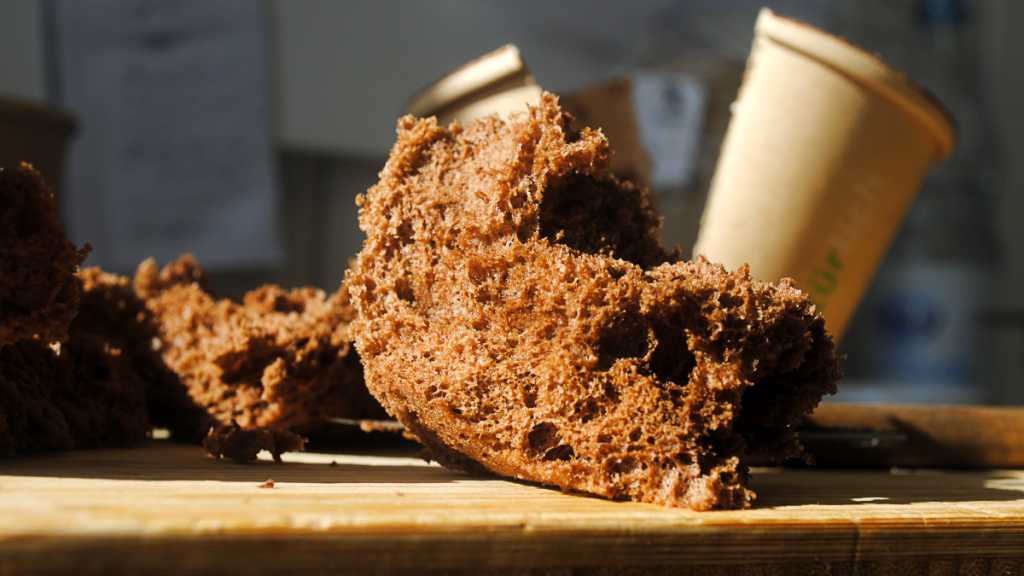 Aufgeschnittener Schokoladen-Sponge mit luftig-poröser Krume auf Holzbrett, im Hintergrund Coffee-to-go-Becher
